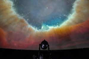 Un proyector de estrellas Zeiss Modelo IX frente a la Nebulosa Helix proyectado sobre la cúpula. Mientras que los proyectores de estrellas crean un cielo nocturno realista, los modernos sistemas digitales de domo completo permiten viajar a través del espacio y el tiempo, a galaxias distantes y mundos alienígenas, pero también al amanecer de los tiempos.